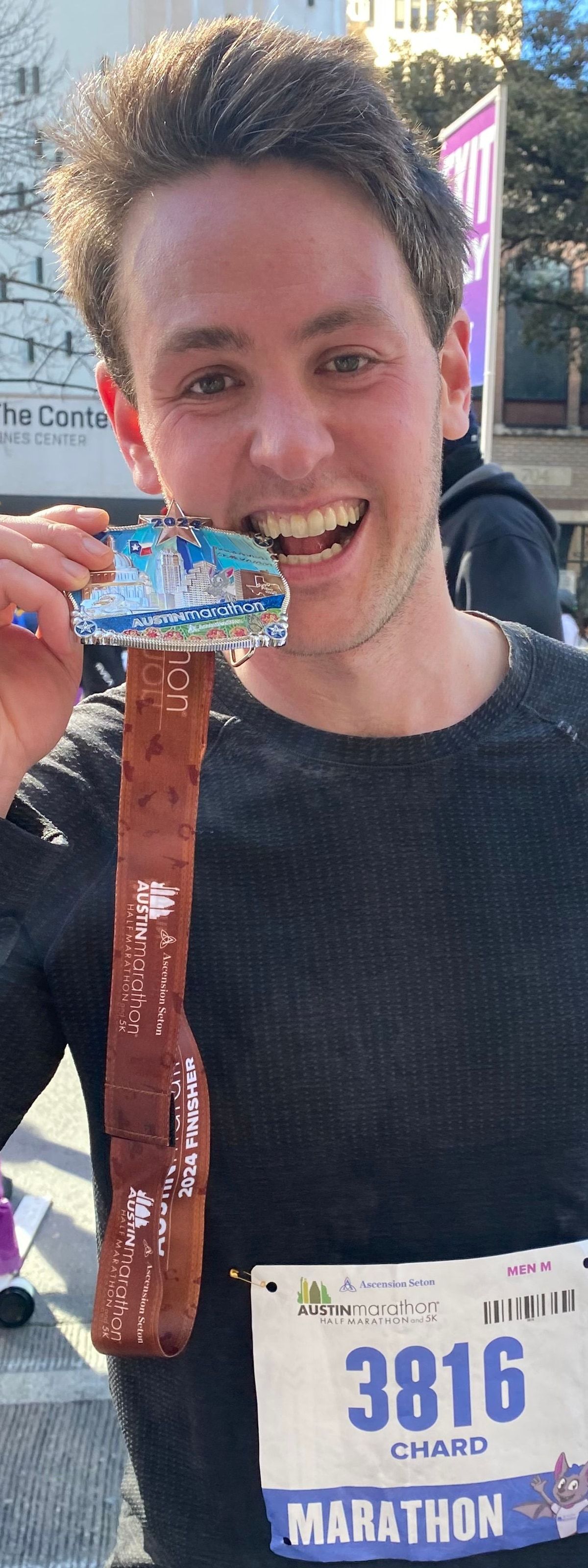 Richard Joseph Spinelli grinning at the 2024 Austin Marathon finish, his medal held up by its strap and bib 3816 on his shirt
