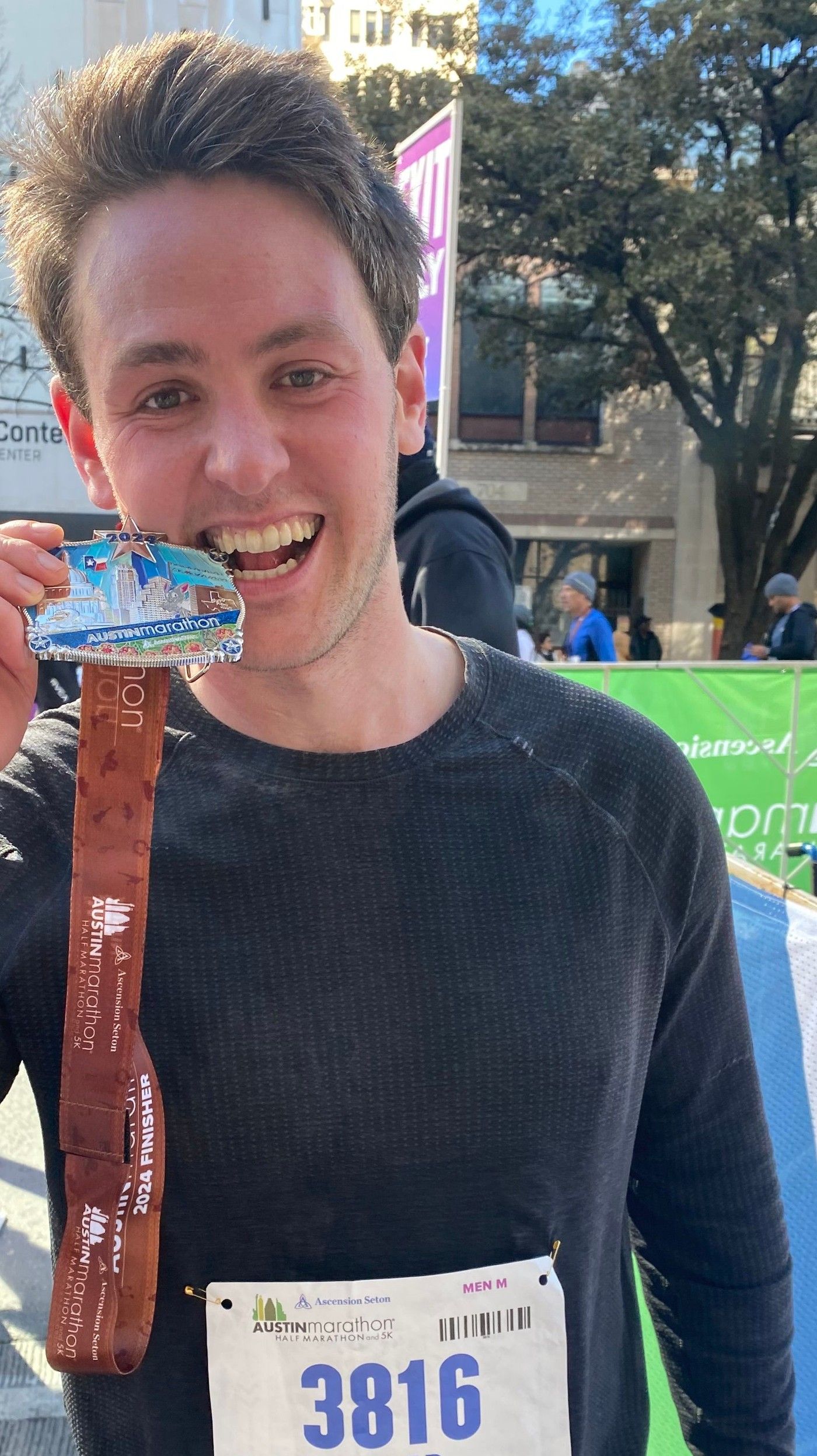Richard Joseph Spinelli holding his finisher medal up to the camera after the 2024 Austin Marathon, bib number 3816 pinned to his shirt